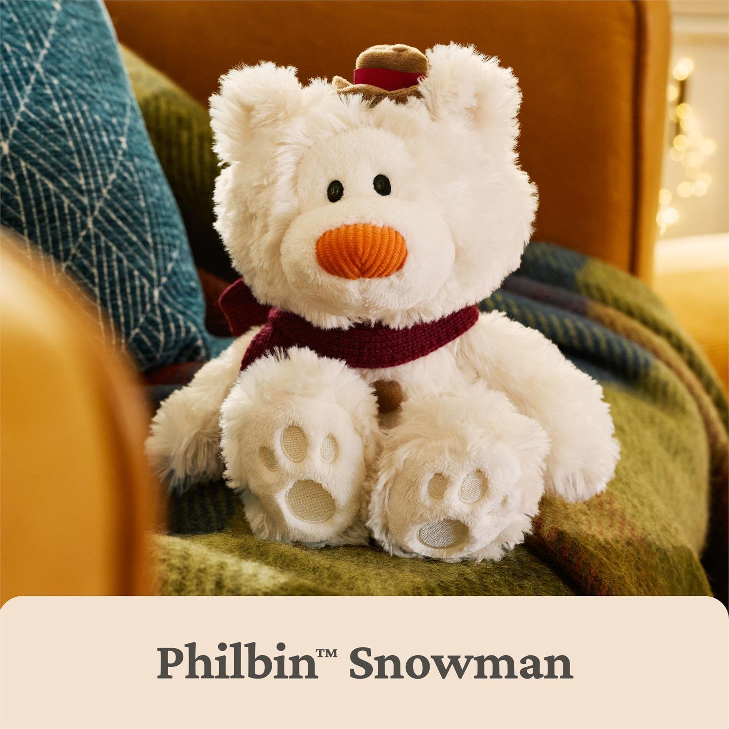 A plush teddy bear styled as a snowman, featuring white faux fur, a dark red scarf, an orange textured nose, and a small brown hat, sitting on a cozy green blanket in a warm indoor setting. A beige banner at the bottom reads 'Philbin Snowman'.