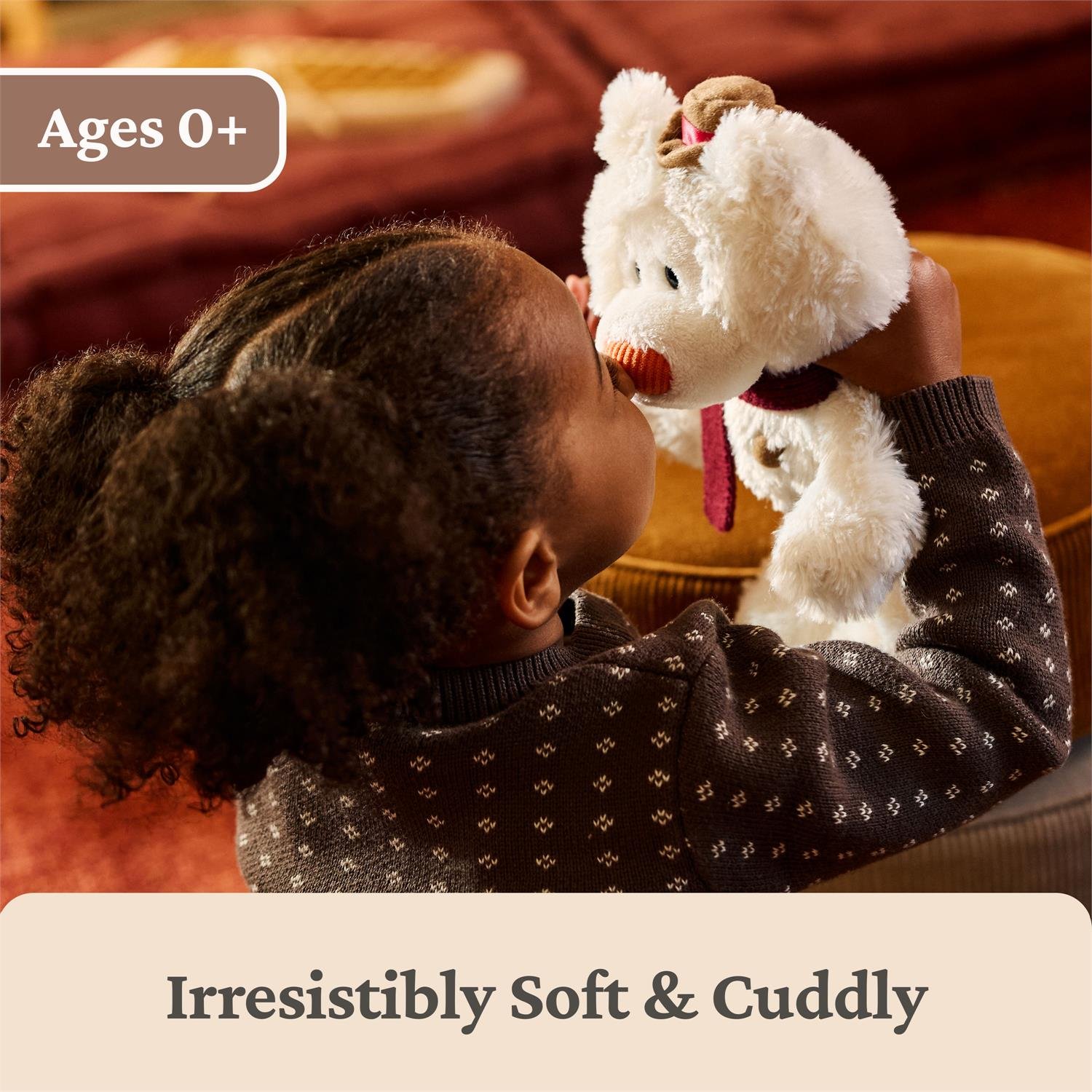A child in a dark patterned sweater holds and kisses a fluffy white stuffed bear with a red nose and red scarf in a cozy indoor setting. Text overlays read 'Ages 0+' and 'Irresistibly Soft & Cuddly'.