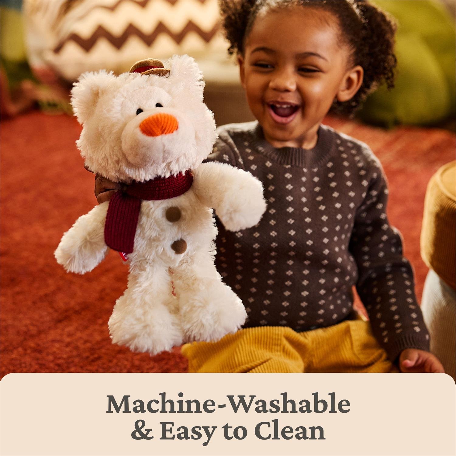 A smiling young child sits on a red carpet holding a plush cream teddy bear with an orange carrot nose, brown buttons, and a dark red scarf. The child wears a dark patterned sweater and mustard yellow pants.
