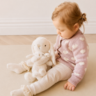 Cream plush hugging bunny set by Snuggle Bunnies, mama and baby, soft textured fur, side view with child in pink heart cardigan, beige ribbed pants, ruffled socks, sitting on neutral rug.