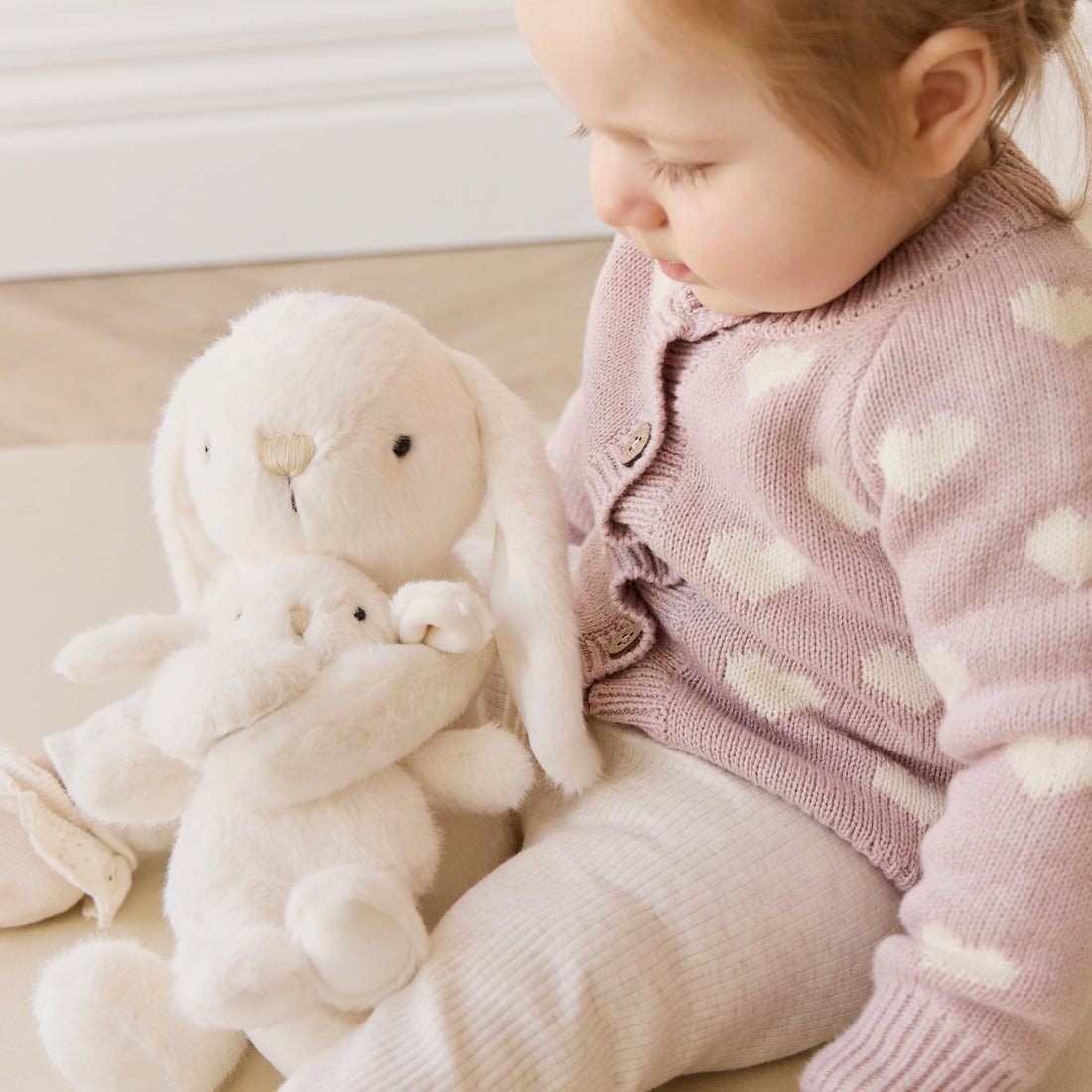 Child in a pink sweater with a plush toy on a light wooden floor.