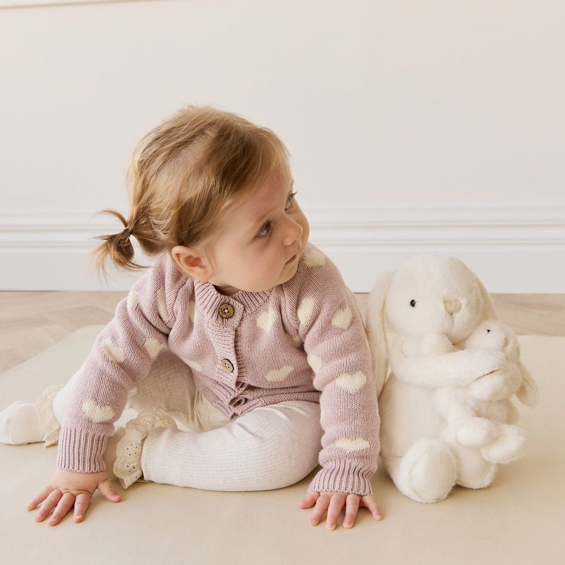 Child in a polka dot cardigan sitting on a light-colored floor with a white teddy bear.