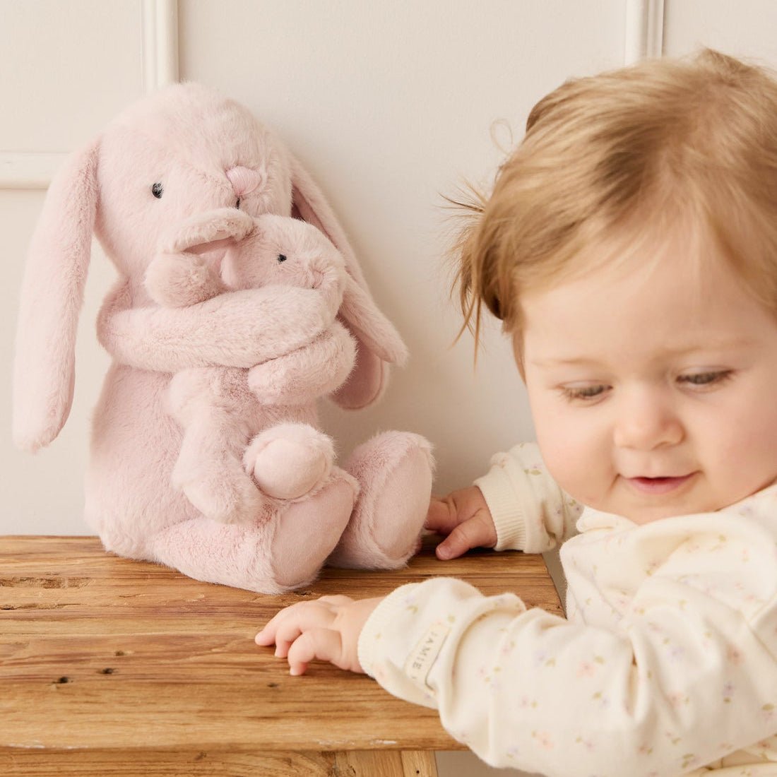 A baby reaching for a pink stuffed bunny on a wooden table.