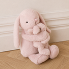 Soft pink plush bunny hugging baby bunny, sitting on wooden floor, side view, cuddly texture, long floppy ears, black button eyes