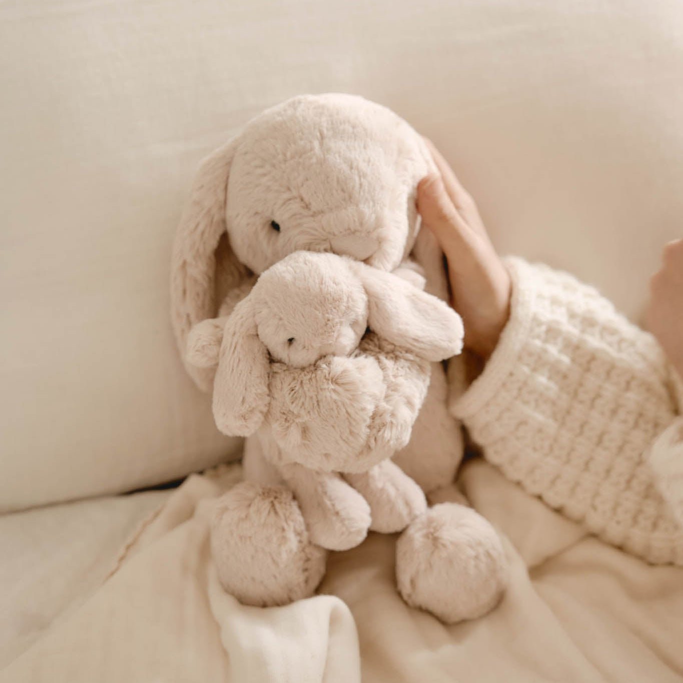 Two beige bunny plush toys stacked on a beige blanket next to a hand wearing a beige knit sweater.