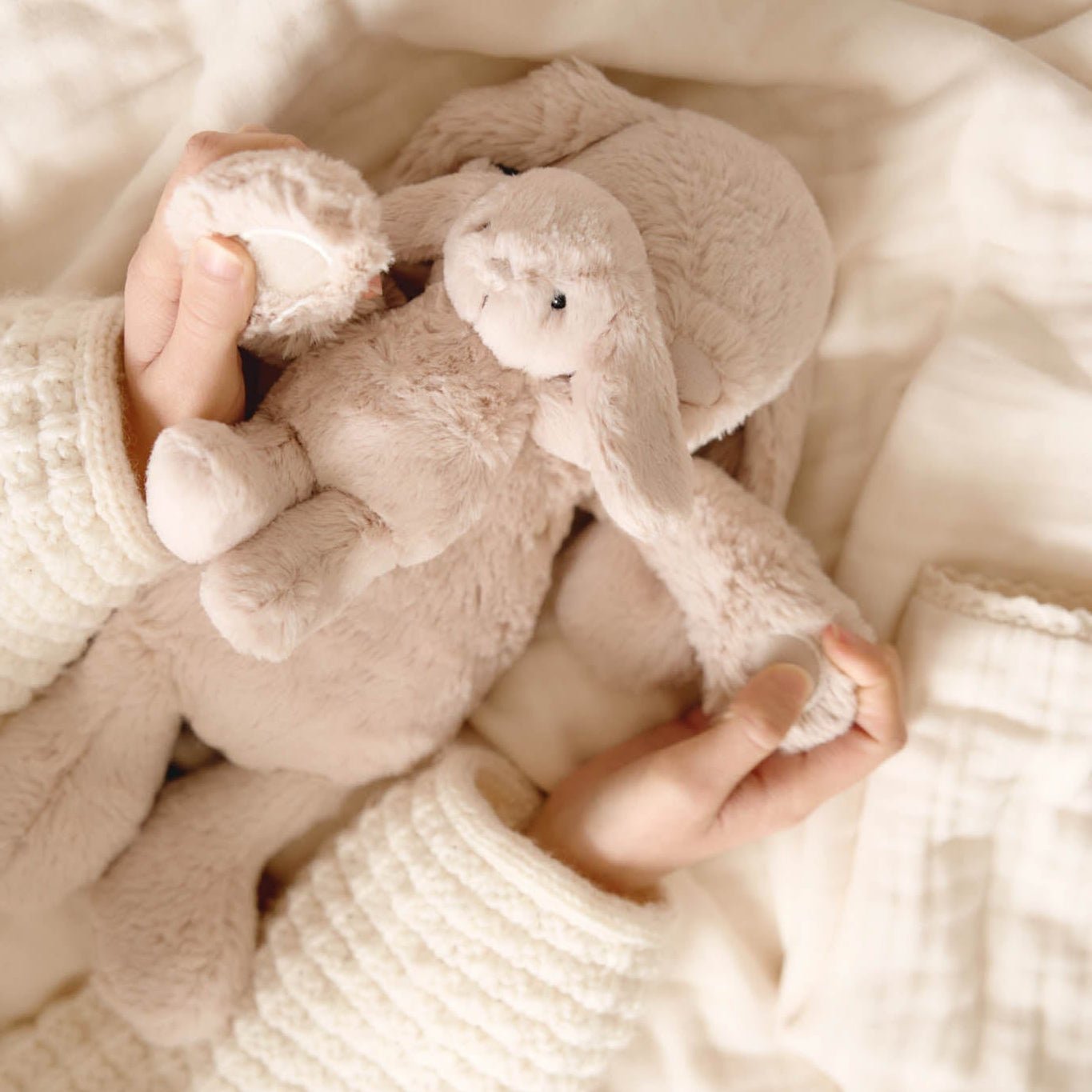 Hands holding stuffed bunny on a white blanket, wearing a cream-colored knit sweater.