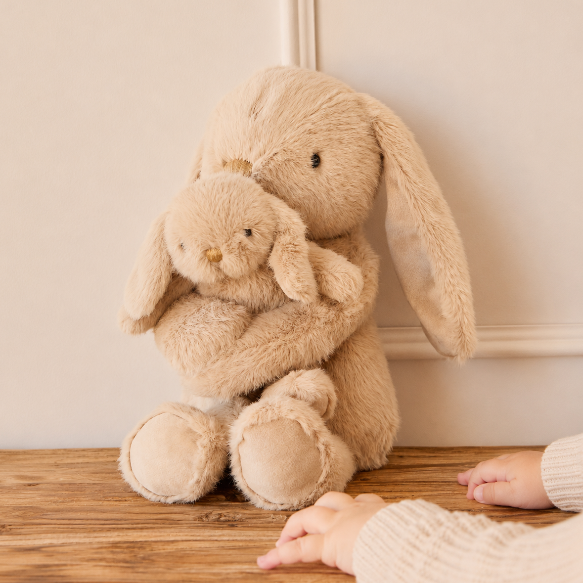 Soft beige plush bunny hugging baby bunny, by Snuggle Bunnies, with long floppy ears, textured fur, and child’s hands nearby on wooden floor.
