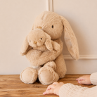 Soft beige plush bunny hugging baby bunny, by Snuggle Bunnies, with long floppy ears, textured fur, and child’s hands nearby on wooden floor.